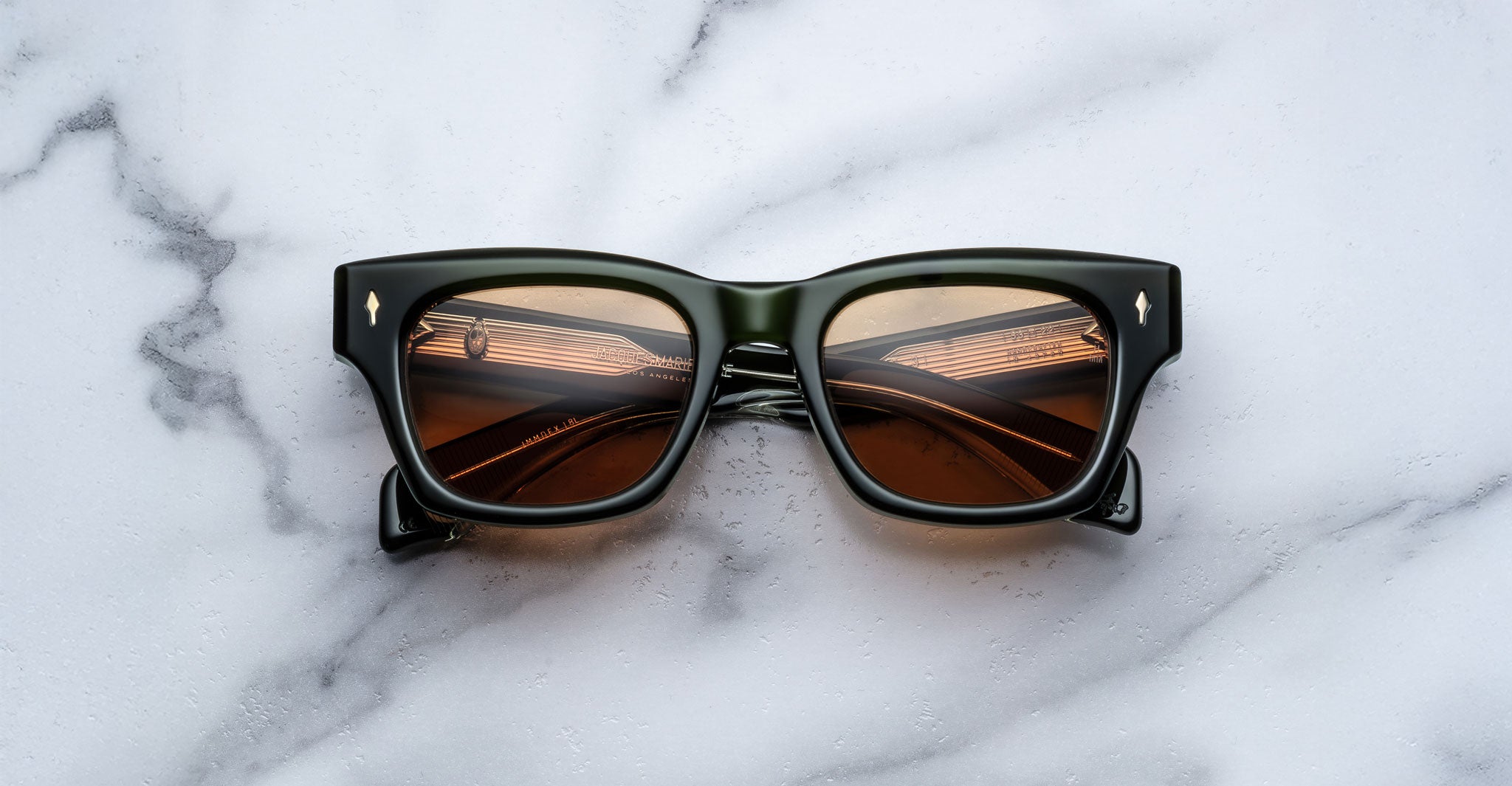 A pair of Jacques Marie Mage Dealan 53 dark square sunglasses with brown lenses rests folded on a white marble surface, showing reflections in the lenses.