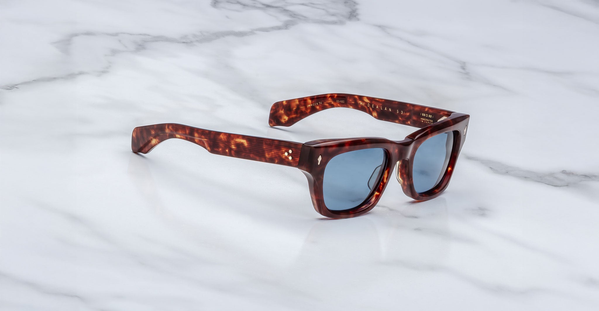 The Jacques Marie Mage Dealan 53 sunglasses, featuring thick tortoiseshell-patterned frames and dark lenses, are placed on a white marble surface with gray veining.