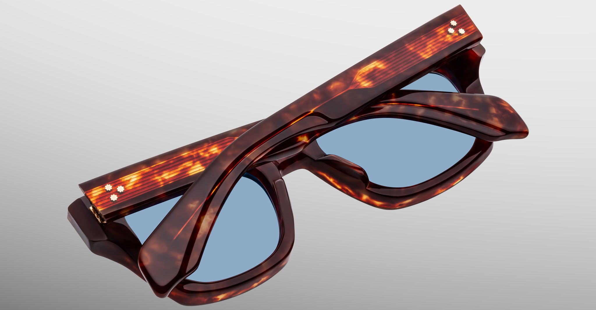 The Jacques Marie Mage Dealan 53 sunglasses, featuring a tortoiseshell pattern and blue lenses, are shown folded against a neutral gradient background.