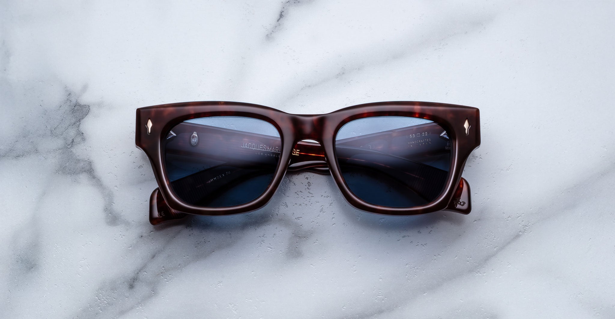 The Jacques Marie Mage Dealan 53, a pair of square dark brown tortoiseshell sunglasses with dark lenses, is displayed on a white marble surface.