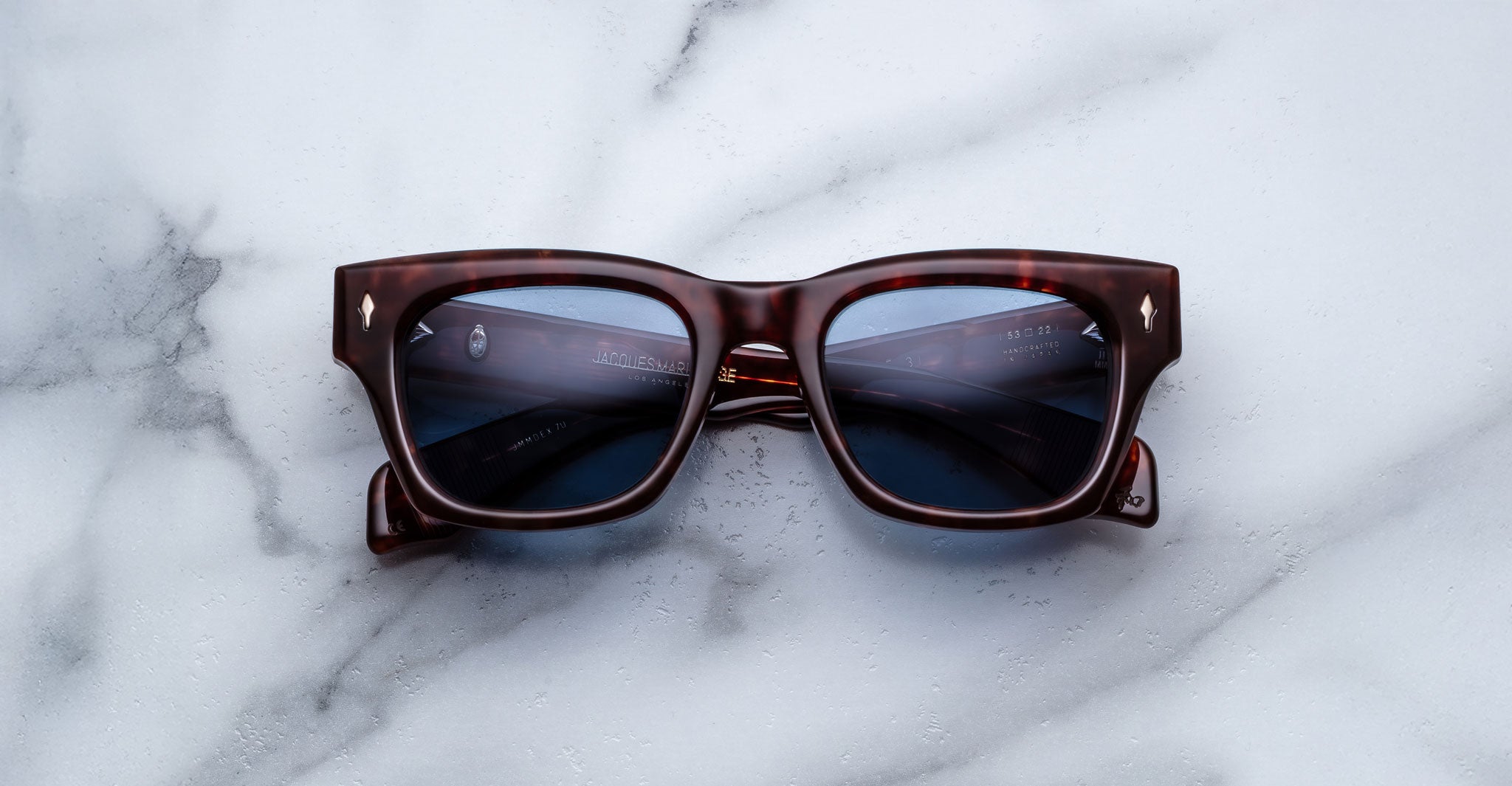 The Jacques Marie Mage Dealan 53, a pair of square dark brown tortoiseshell sunglasses with dark lenses, is displayed on a white marble surface.