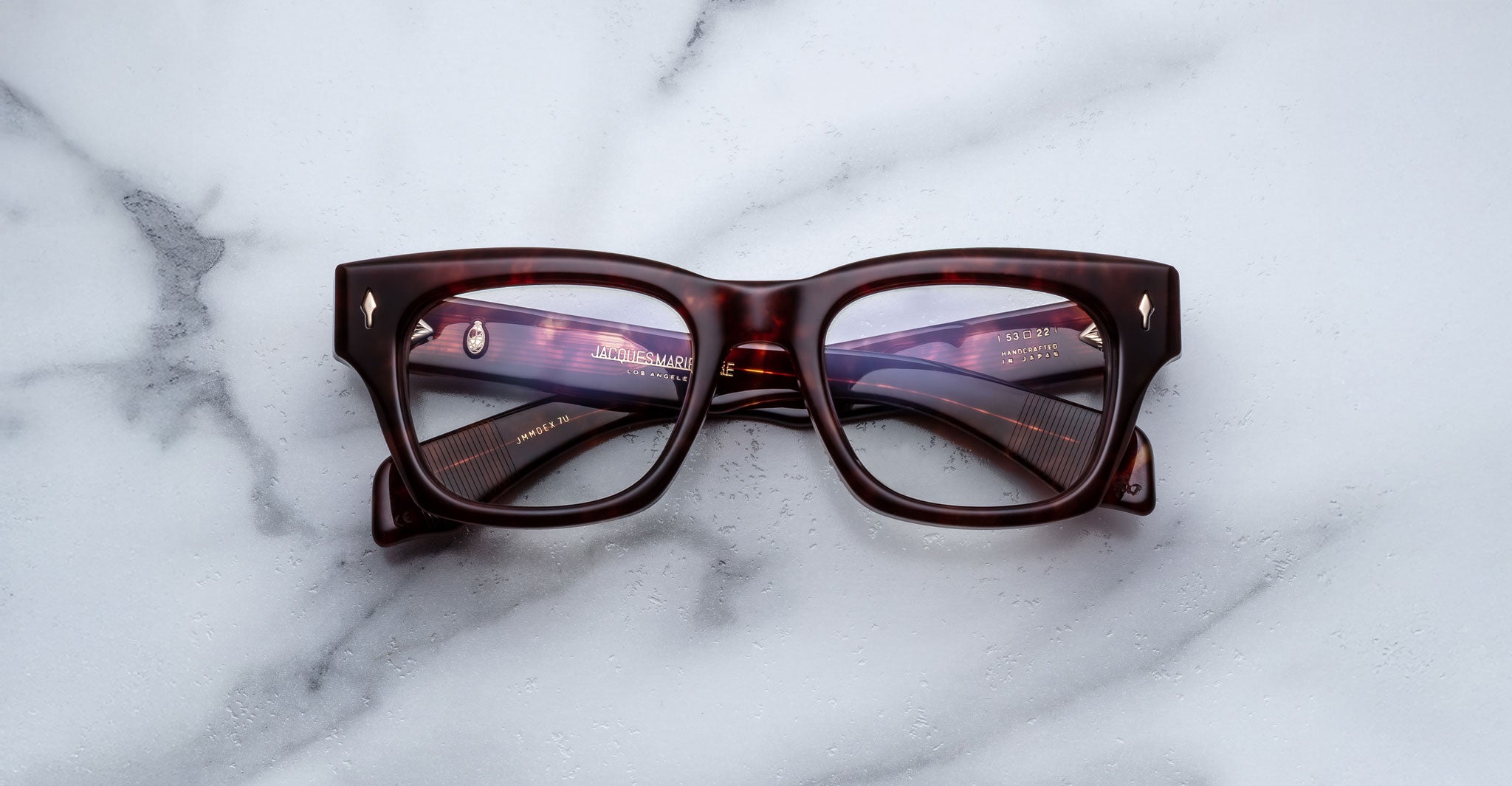 A pair of Jacques Marie Mage Dealan 53 rectangular eyeglasses with thick, dark tortoiseshell frames lies closed on a white marble surface.