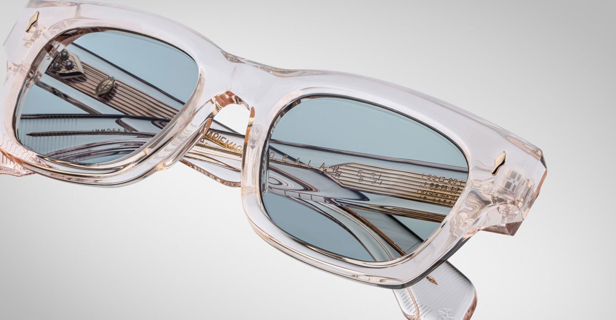 Close-up of Jacques Marie Mage Dealan 53 sunglasses featuring transparent, square frames and blue-tinted lenses, set against a plain light gray background with metallic reflections visible in the lenses.