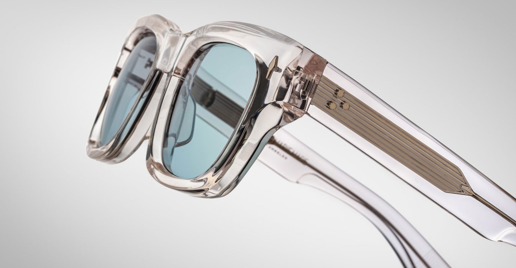 Close-up of Jacques Marie Mage Dealan 53 sunglasses featuring thick, clear frames with blue-tinted lenses and visible metallic details inside the arms, set against a plain light gray background.