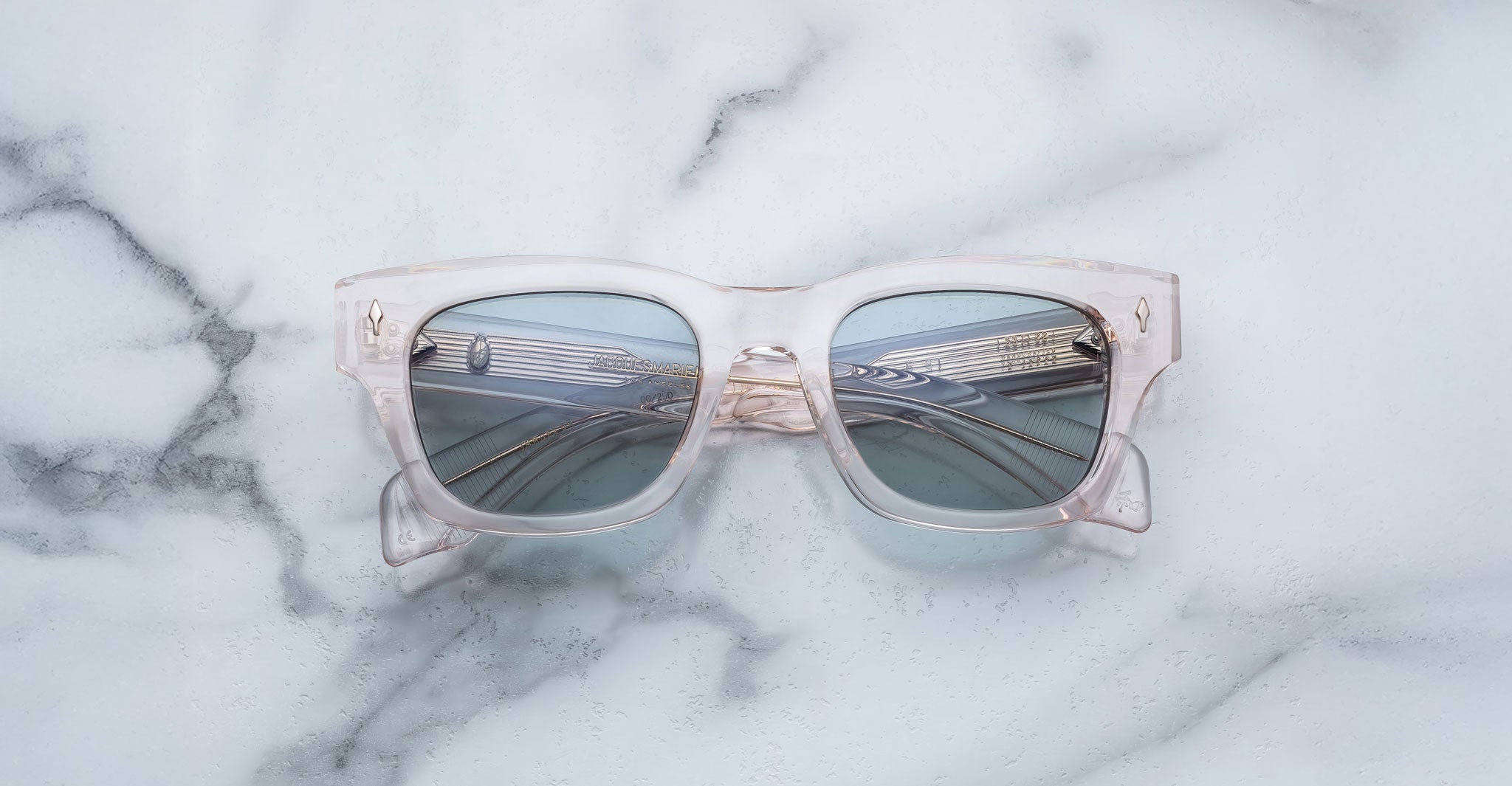 A pair of Jacques Marie Mage Dealan 53 clear, square-framed sunglasses with blue-tinted lenses rests on a white marble surface, the lenses reflecting a metal rail structure.