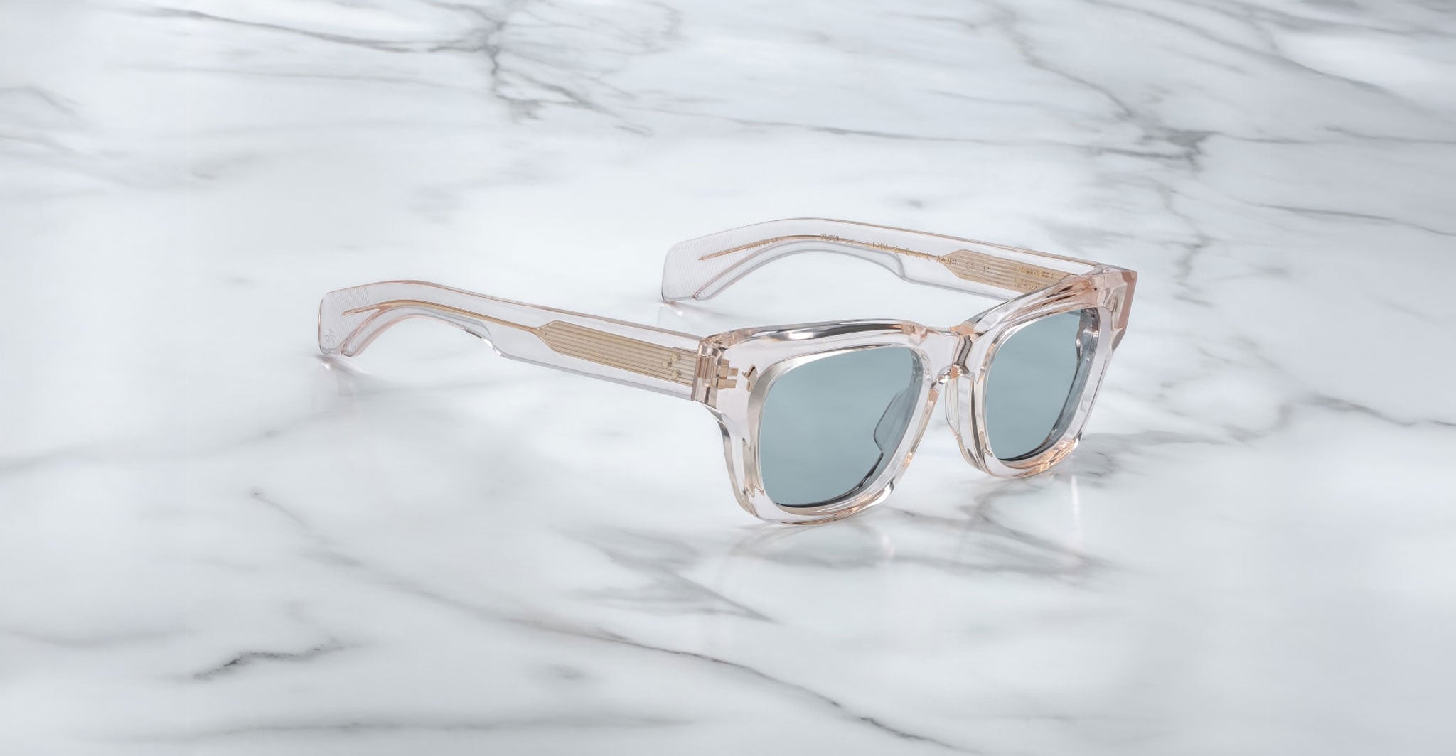 The Jacques Marie Mage Dealan 53 sunglasses, featuring clear frames and light blue-tinted lenses, rest on a white marble surface with grey veining. Gold accents highlight the arms near the hinges.