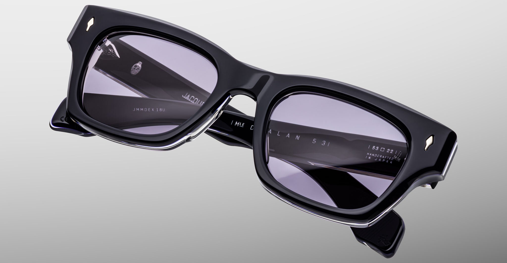 A close-up of Jacques Marie Mage Dealan 53 sunglasses showcases thick black rectangular frames, tinted lenses, and detailed branding and hinge reflections on a light gray background.