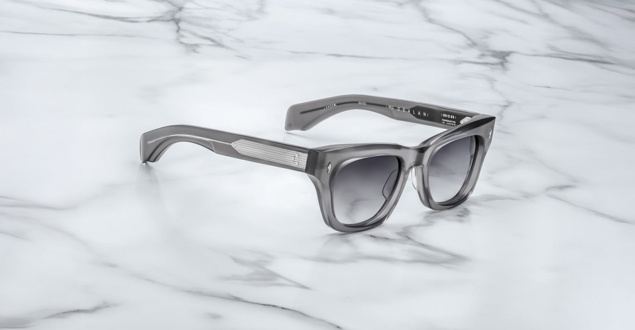 The Jacques Marie Mage Dealan 53 sunglasses, featuring gray thick frames and dark gradient lenses, rest on a white marble surface with gray veining.