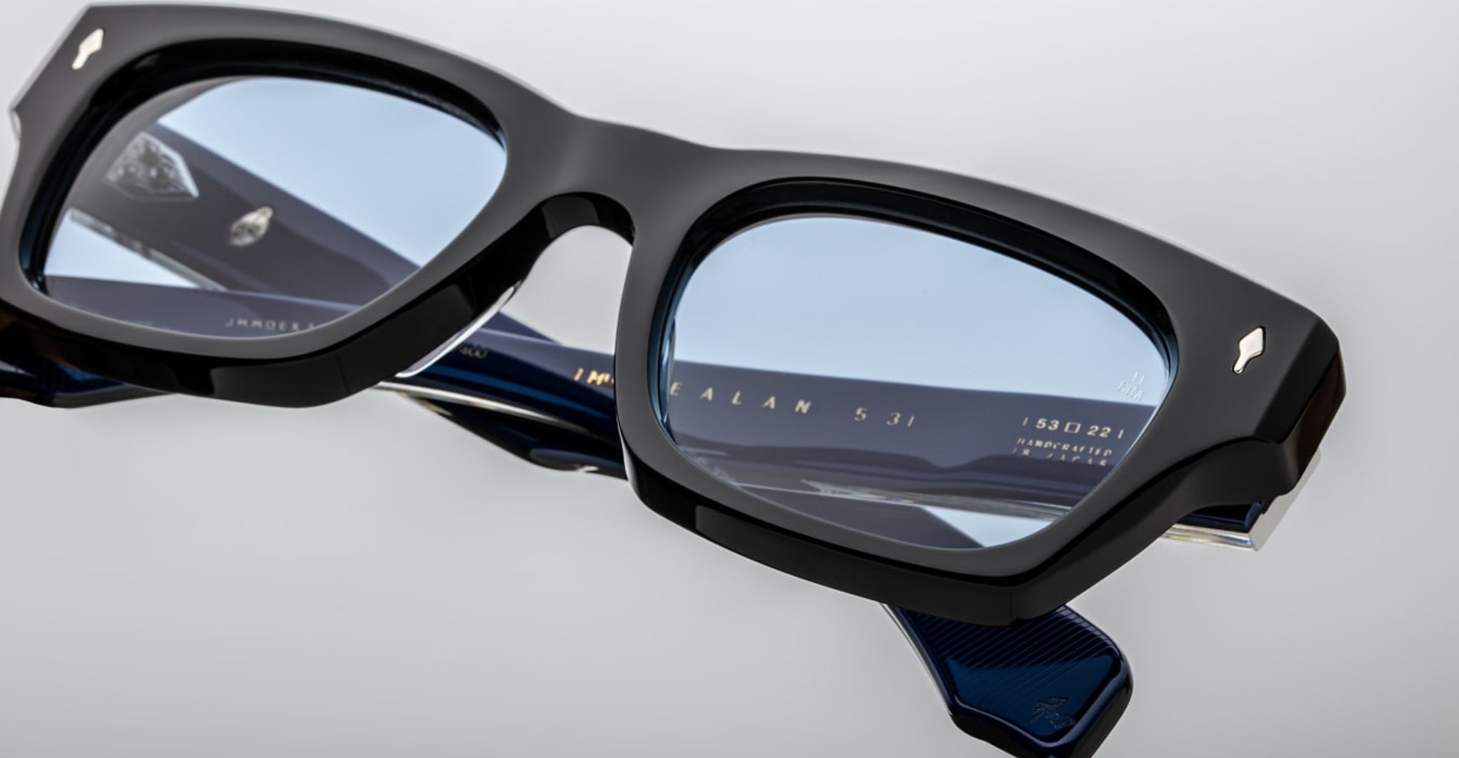 A close-up of Jacques Marie Mage Dealan 53 rectangular eyeglasses with thick black frames and blue-tinted lenses, resting on a flat surface. Gold text appears on the temple arms.