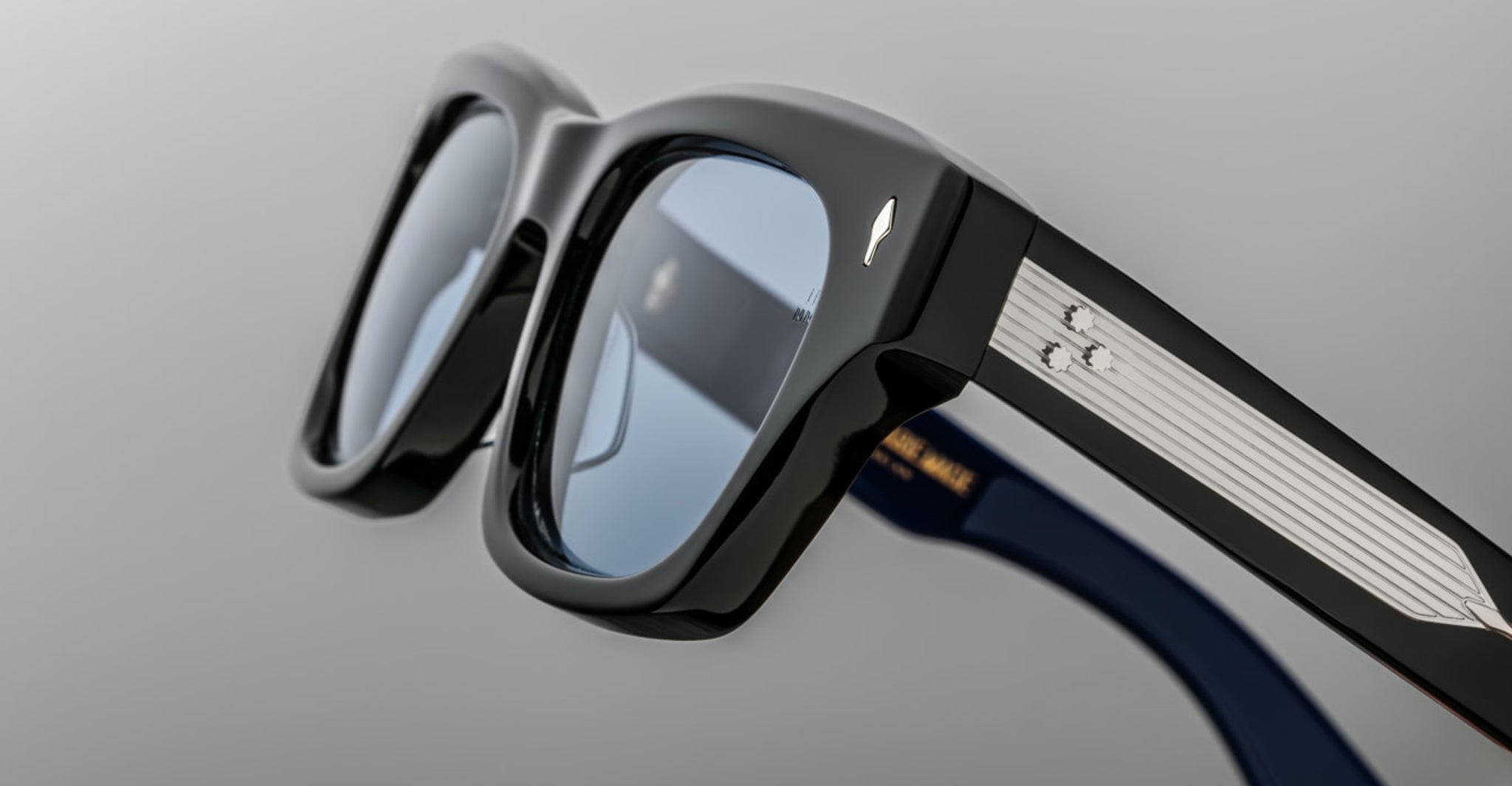 Close-up of Jacques Marie Mage Dealan 53 black rectangular eyeglass frames with blue-tinted lenses, silver detailing on the arms, a subtle side logo, and a neutral gray background.