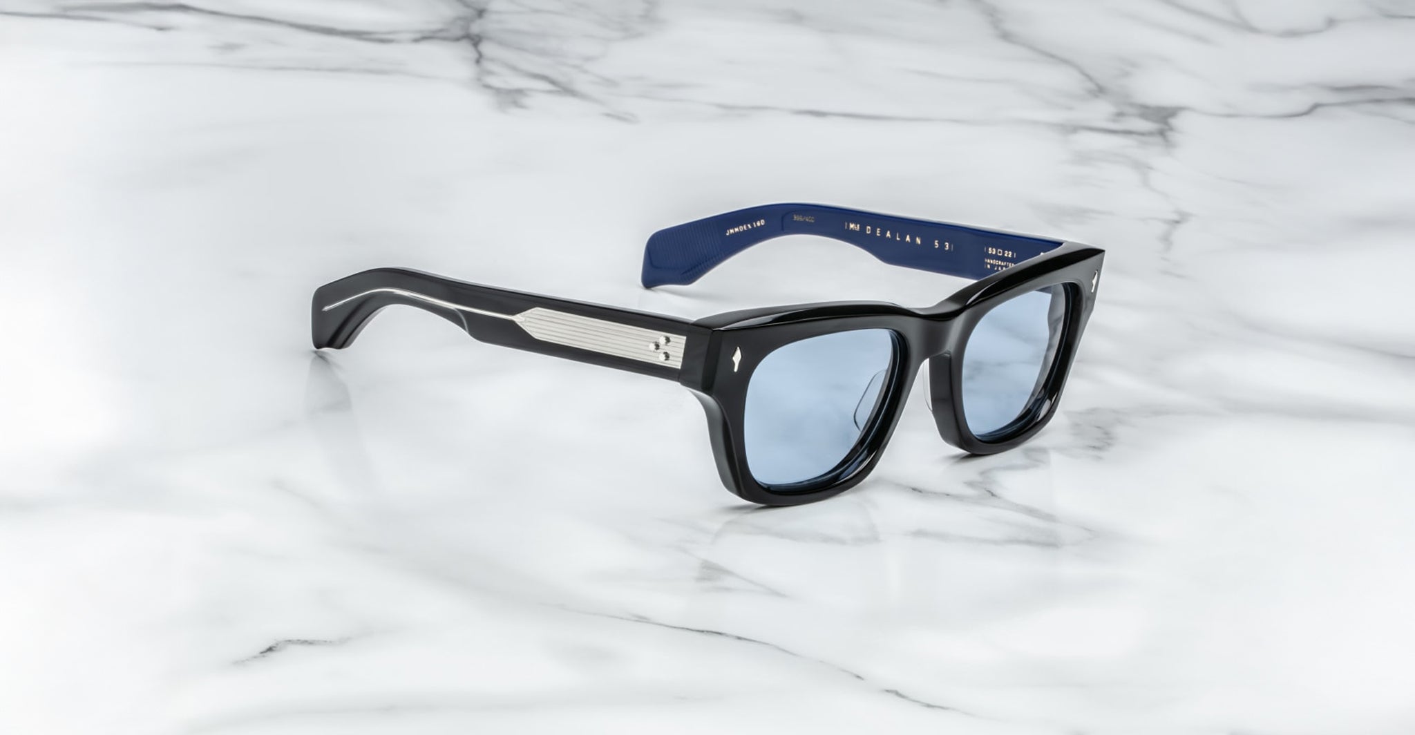 The Jacques Marie Mage Dealan 53 sunglasses, featuring a black rectangular frame, light blue lenses, and metal temple accents, are displayed on a white marble surface.