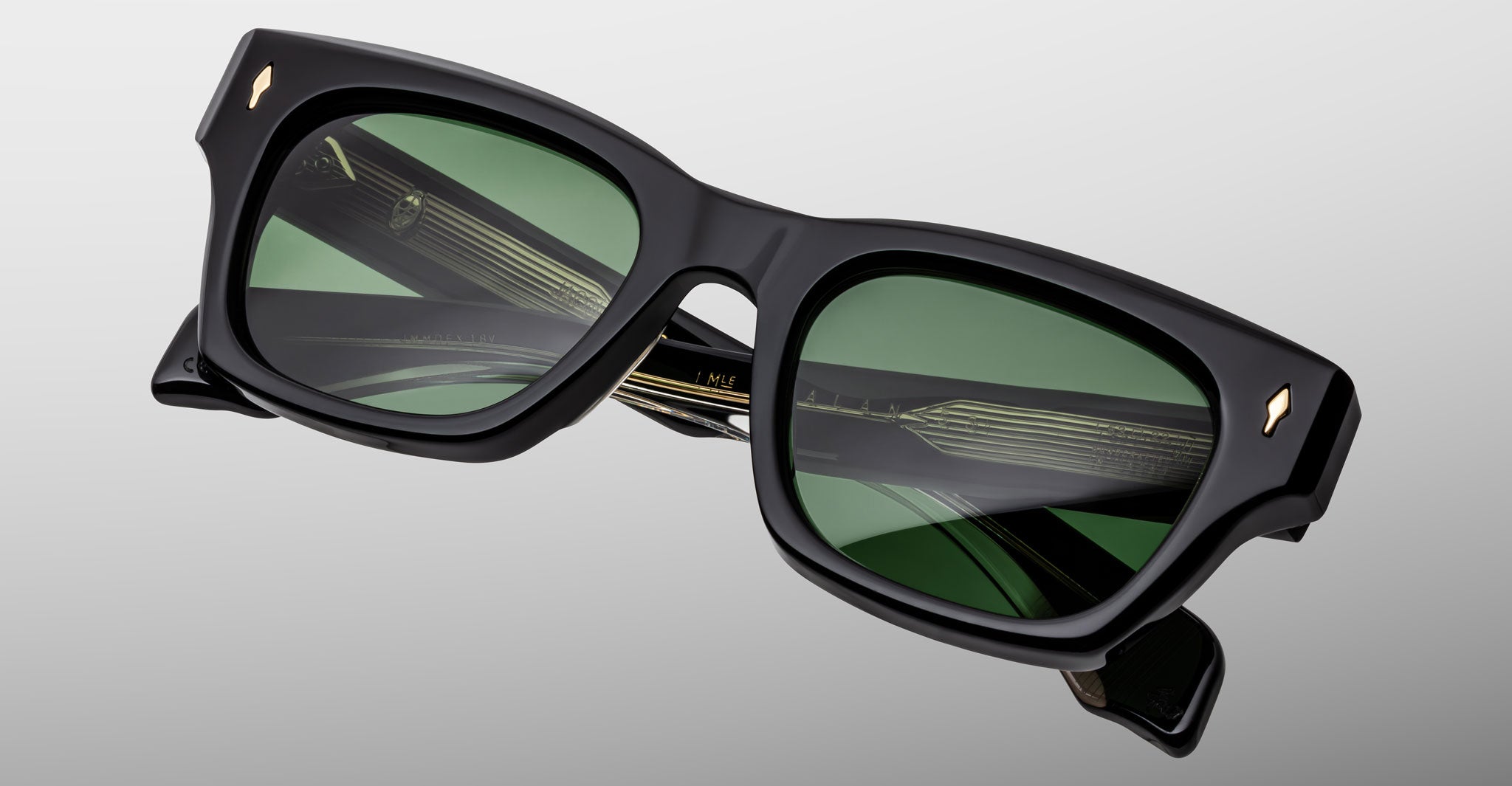 A close-up of Jacques Marie Mage Dealan 53 sunglasses, featuring a black rectangular frame, dark green lenses, and gold hinge accents, set against a plain light gray background.