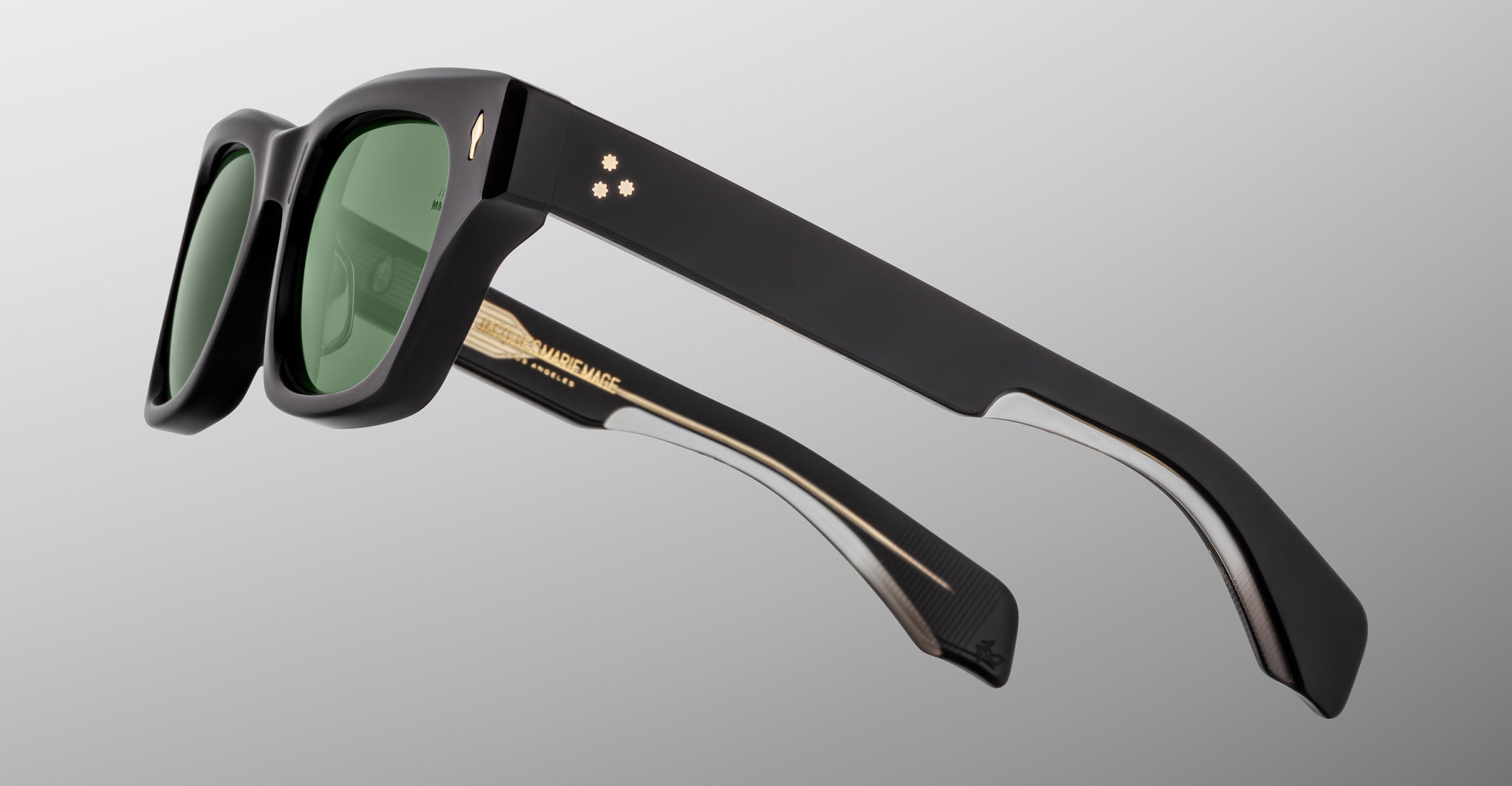 The Jacques Marie Mage Dealan 53 sunglasses feature a black rectangular frame, green lenses, gold accents, and two small gold dots on the temple, shown against a light grey gradient background.