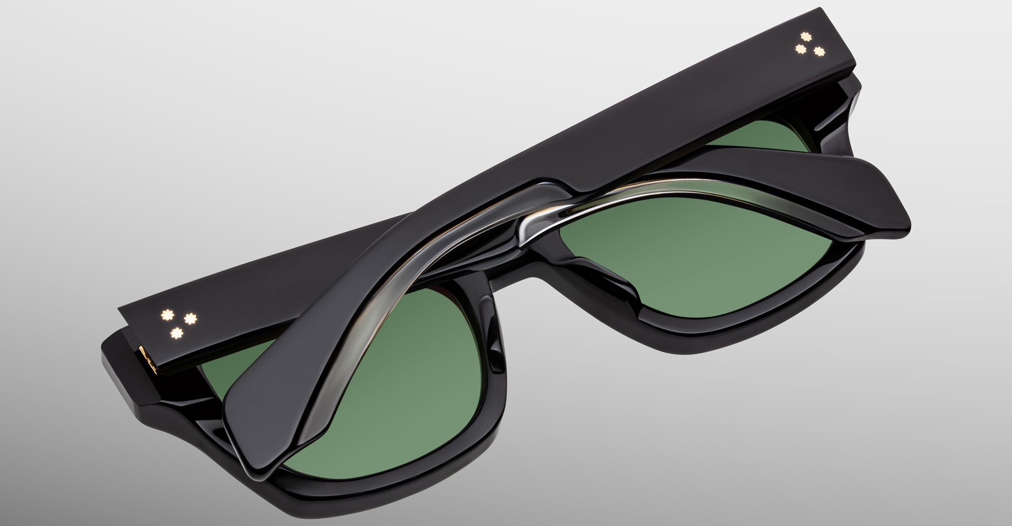 The Jacques Marie Mage Dealan 53, a pair of black sunglasses with green lenses, is displayed folded with the arms crossed neatly over the front against a plain light gray background.