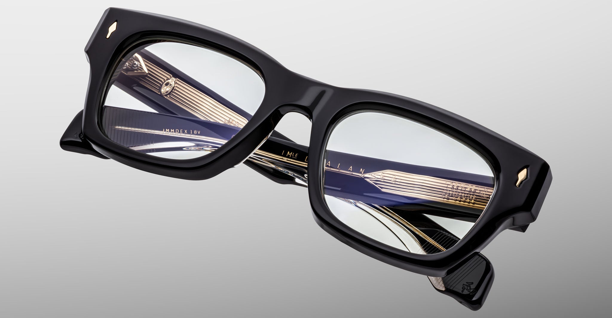 The Jacques Marie Mage Dealan 53 is a pair of black, thick-rimmed eyeglasses with decorative gold and black detailing on the inner arms, displayed against a light gray background.