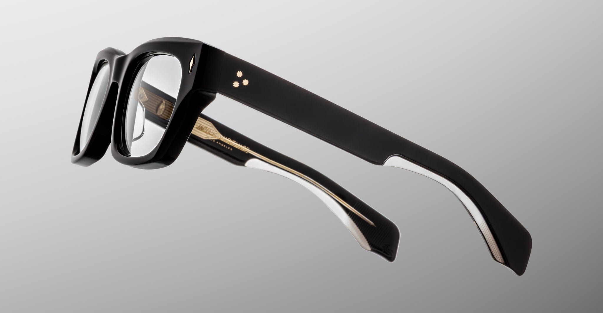 The Jacques Marie Mage Dealan 53 black rectangular eyeglasses feature thick frames, gold contacts by the left hinge, and a white accent along the inner arms, displayed from the side against a plain gray background.