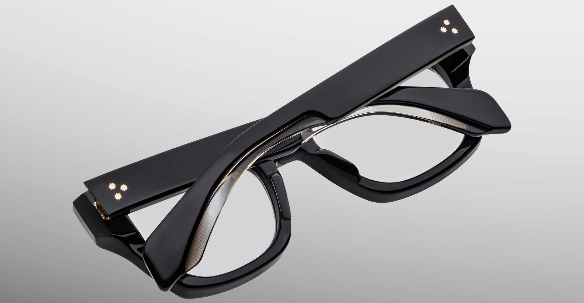 The Jacques Marie Mage Dealan 53 glasses are shown folded closed. They feature thick black frames with small gold-colored circular details on the hinges and temples, set against a light gradient background.