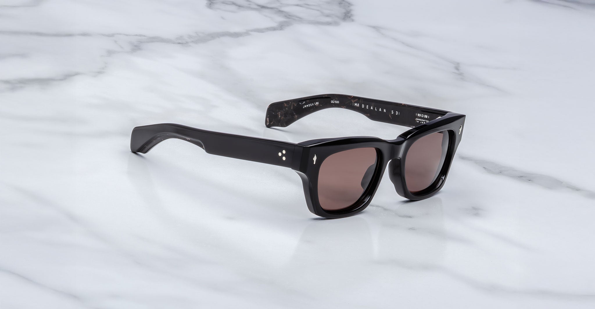 The Jacques Marie Mage Dealan 53 sunglasses, featuring black frames and brown tinted lenses, rest on a white marble surface. The temples show small silver dot accents and text appears on the inside of the right arm.