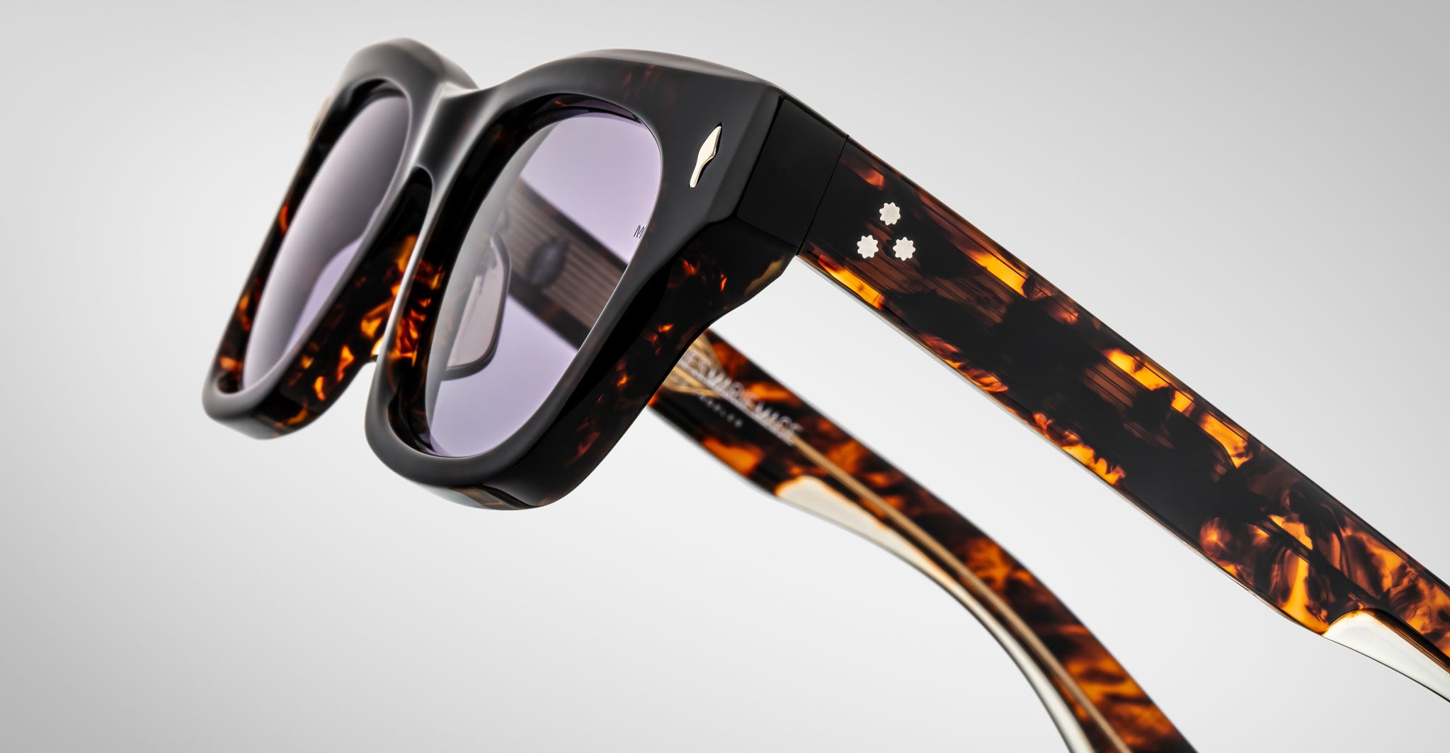 Close-up of Jacques Marie Mage Dealan sunglasses in a tortoiseshell pattern with dark lenses. The frame has two white dot accents and a metallic bird emblem on the arms, set against a light gray background.