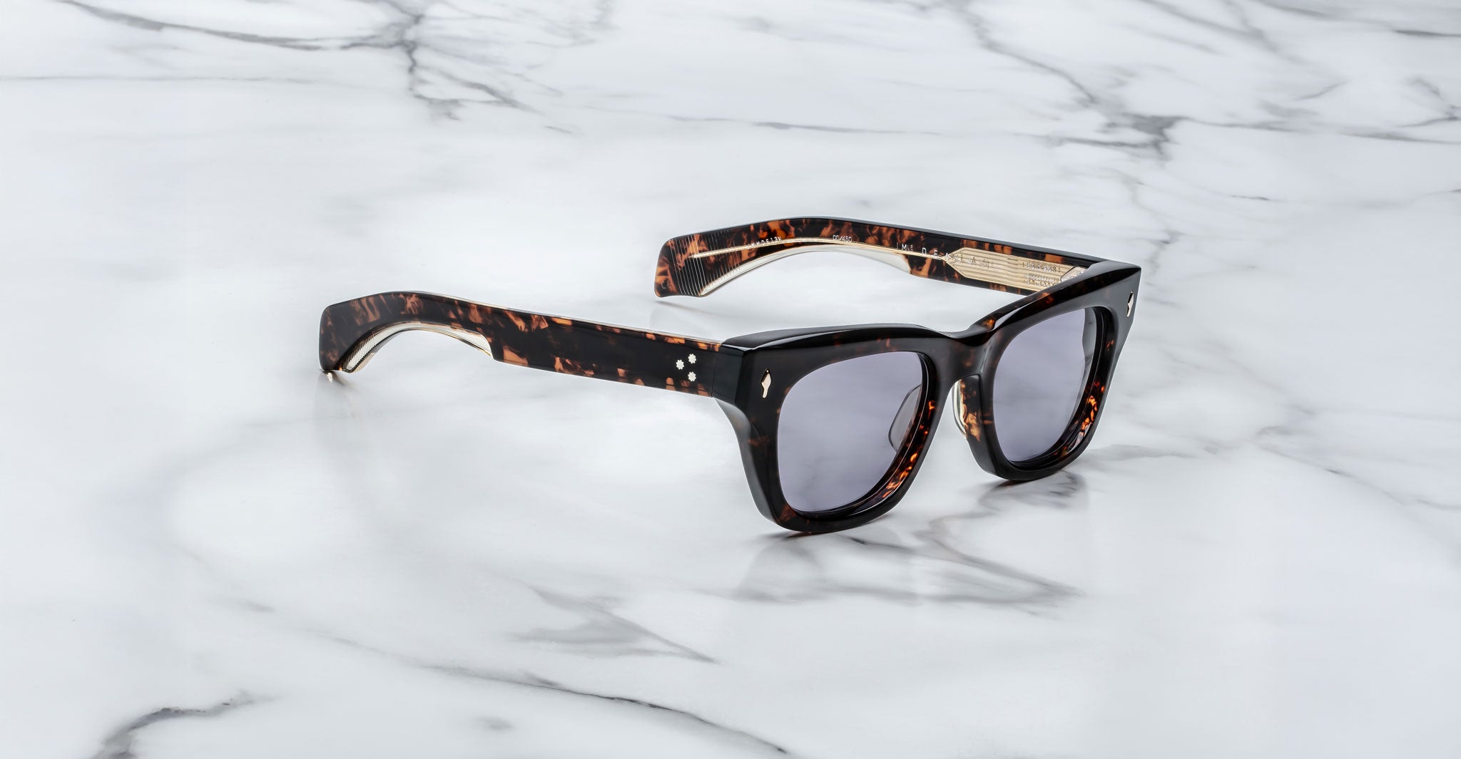 The Jacques Marie Mage Dealan sunglasses, featuring tortoiseshell thick frames and dark lenses, rest on a white marble surface with gray veins.