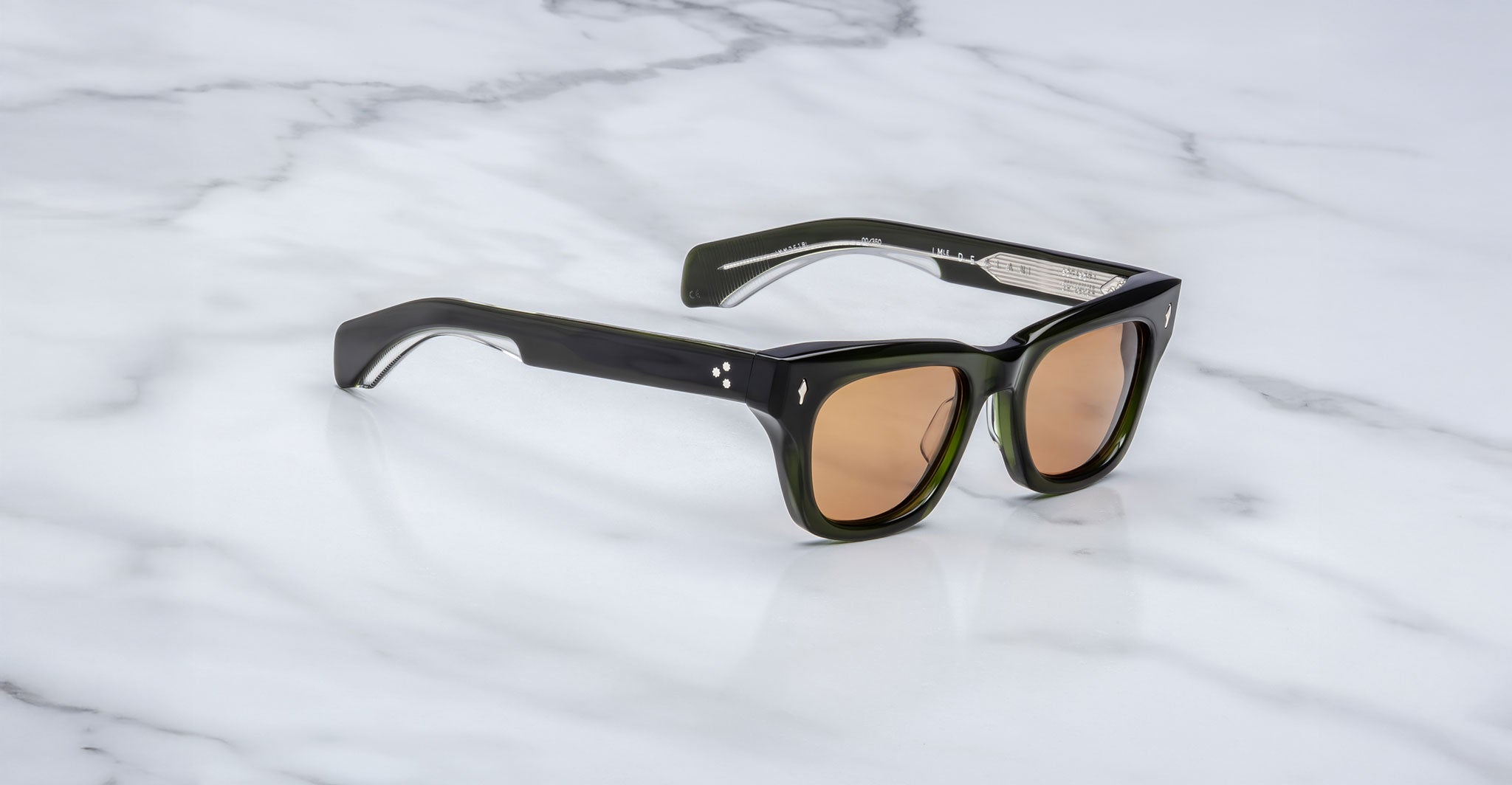The Jacques Marie Mage Dealan sunglasses have thick, glossy black frames with brown-tinted lenses and small metallic details at the hinges, shown resting on a white marble surface.