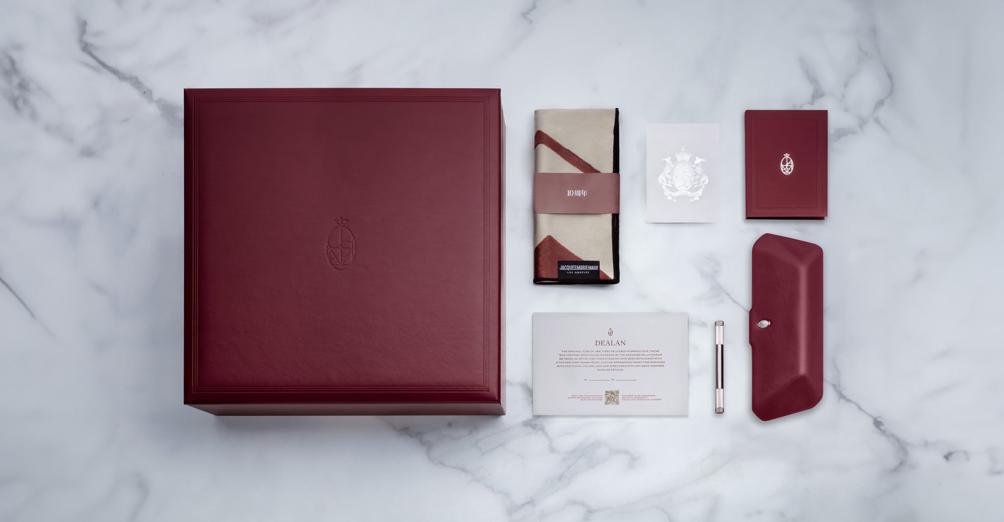 A Jacques Marie Mage Dealan burgundy box with an embossed logo is showcased on marble, accompanied by a folded cloth, certificate, screwdriver, card, matching envelope, and booklet—all neatly arranged.