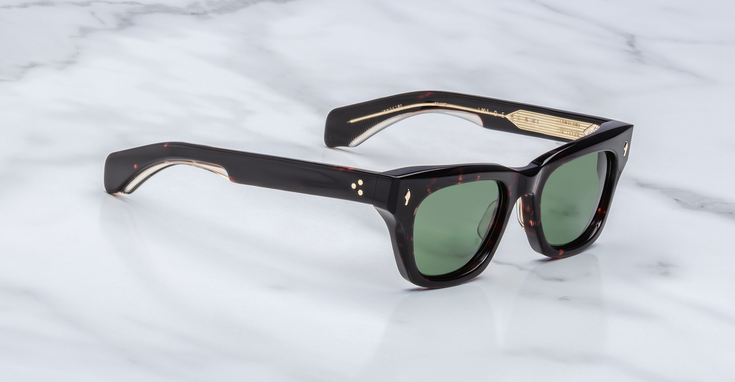 A pair of Jacques Marie Mage Dealan dark tortoiseshell square sunglasses with green lenses and gold accents rests on a white marble surface.