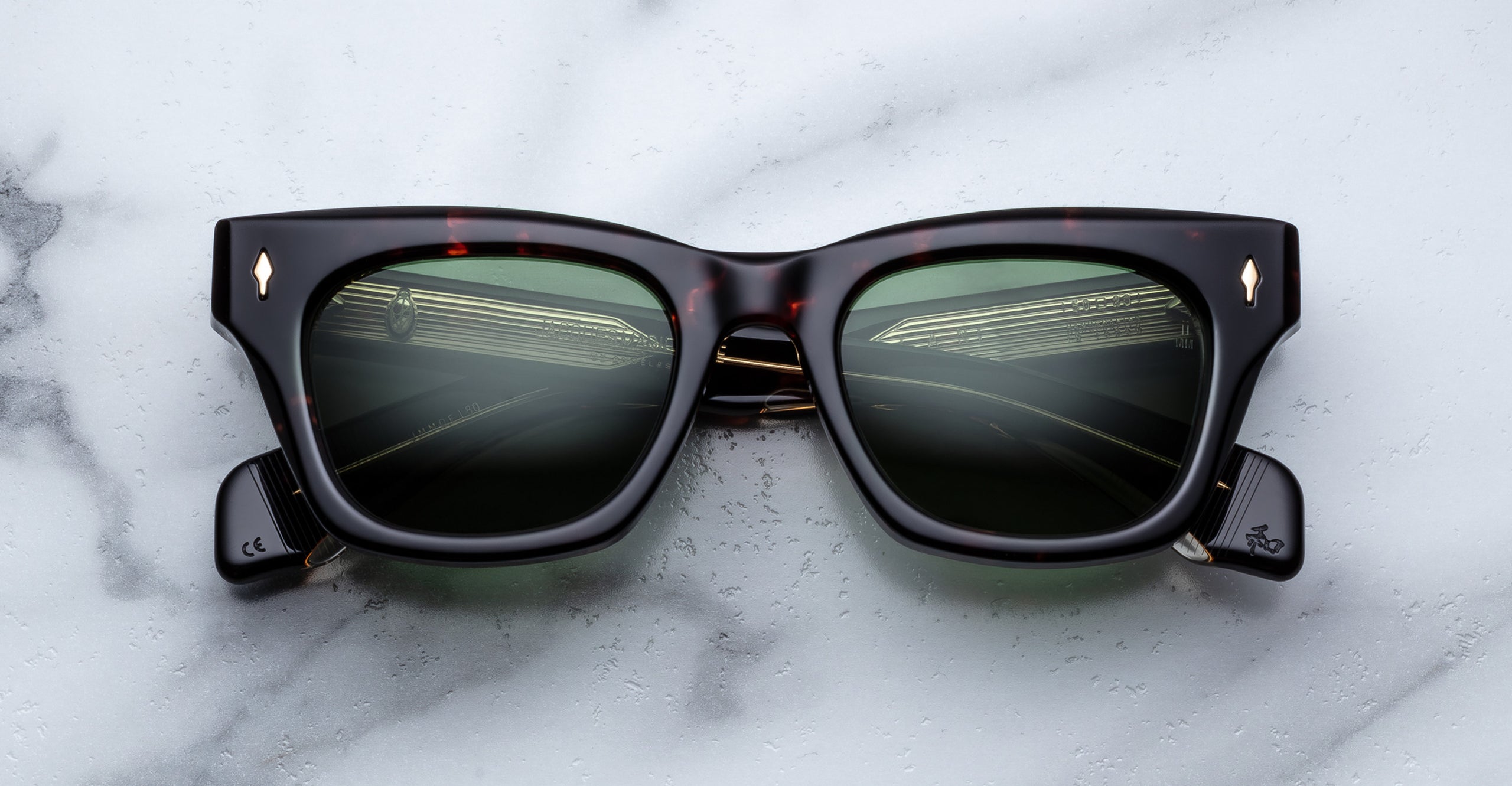 The Jacques Marie Mage Dealan sunglasses, featuring black frames and dark lenses, rest on a white marble surface. The lenses reflect a road with guardrails and a cloudy sky.