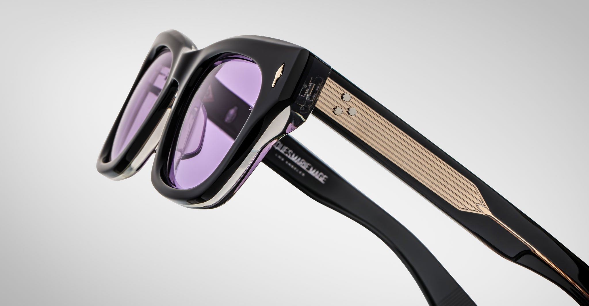 Close-up of Jacques Marie Mage Dealan black sunglasses with thick frames, purple-tinted lenses, and gold accents on the temples, displayed against a plain light gray background.