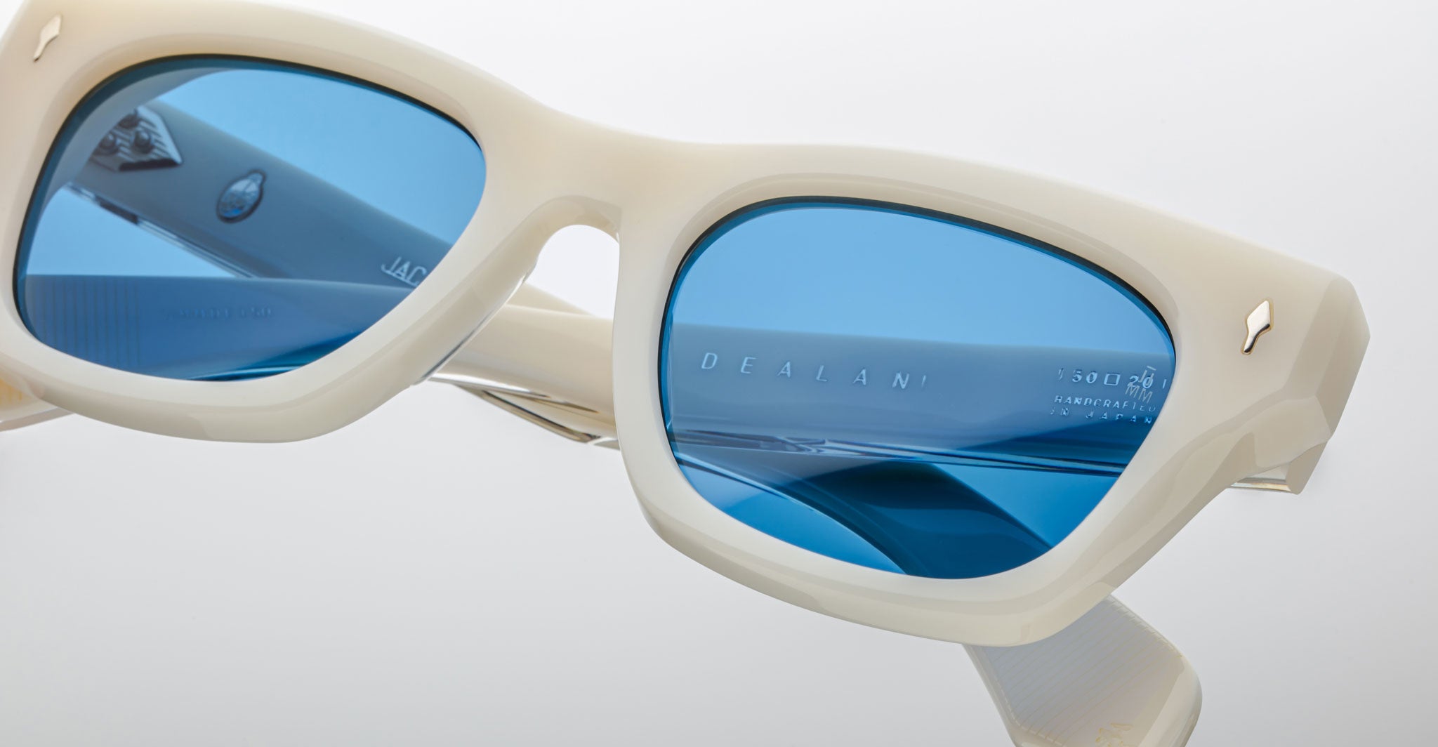 Close-up of cream-colored Jacques Marie Mage Dealan sunglasses with blue tinted lenses, metallic accents on the frame and temple corners, and “DEALAN” visible on the inside of one arm.