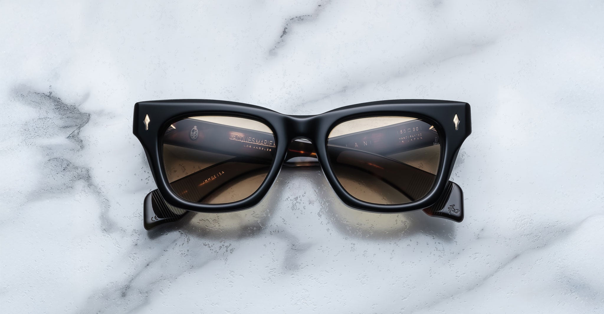 The Jacques Marie Mage Dealan sunglasses feature black square frames with brown-tinted lenses, displayed face up on a white marble surface. Gold writing and small logos embellish the inner arms.