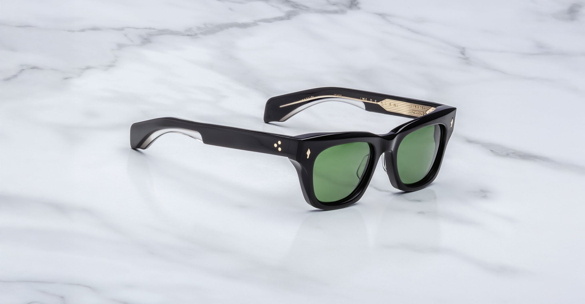 The Jacques Marie Mage Dealan sunglasses, featuring a thick black rectangular frame with a glossy finish, green lenses, and gold-accented arms, rest on a white marble surface.