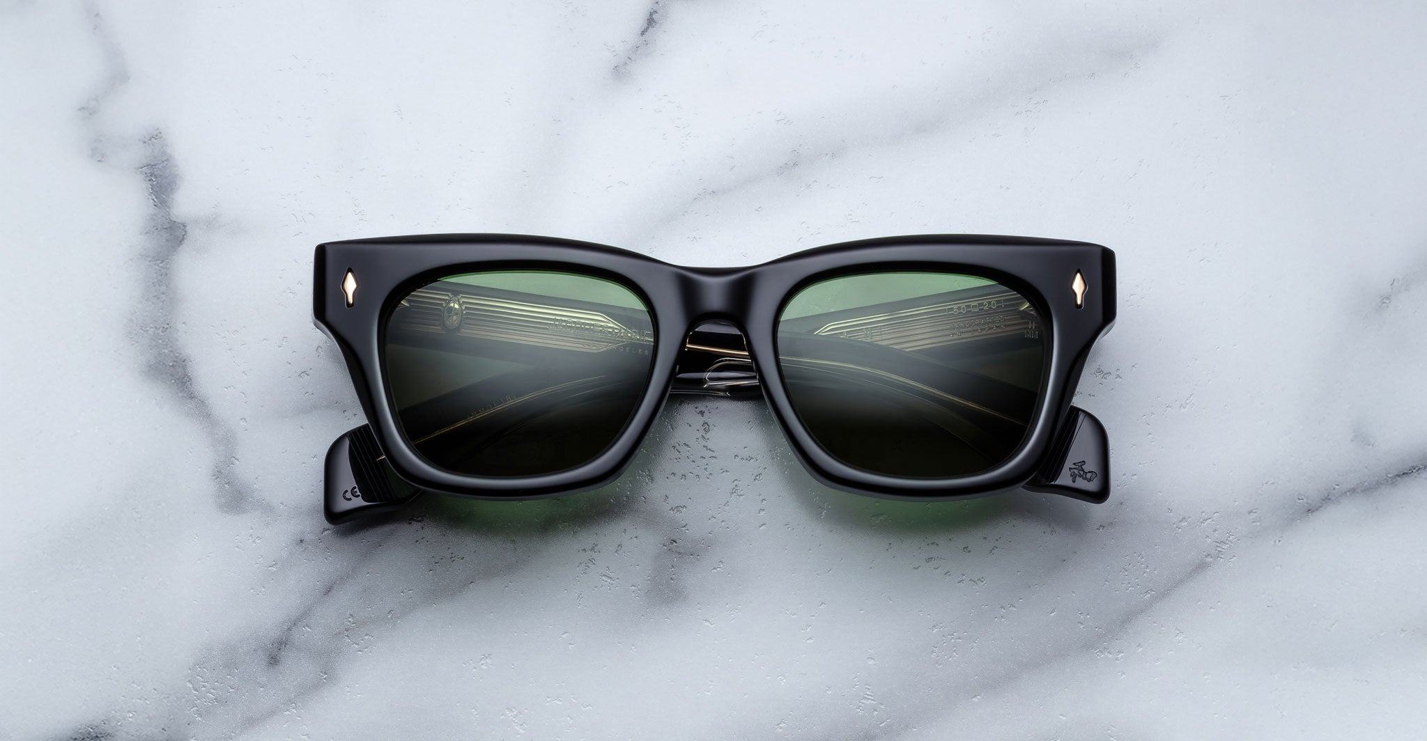 The Jacques Marie Mage Dealan, a pair of black square sunglasses with green-tinted lenses and metal corner accents, rests on a white marble surface.