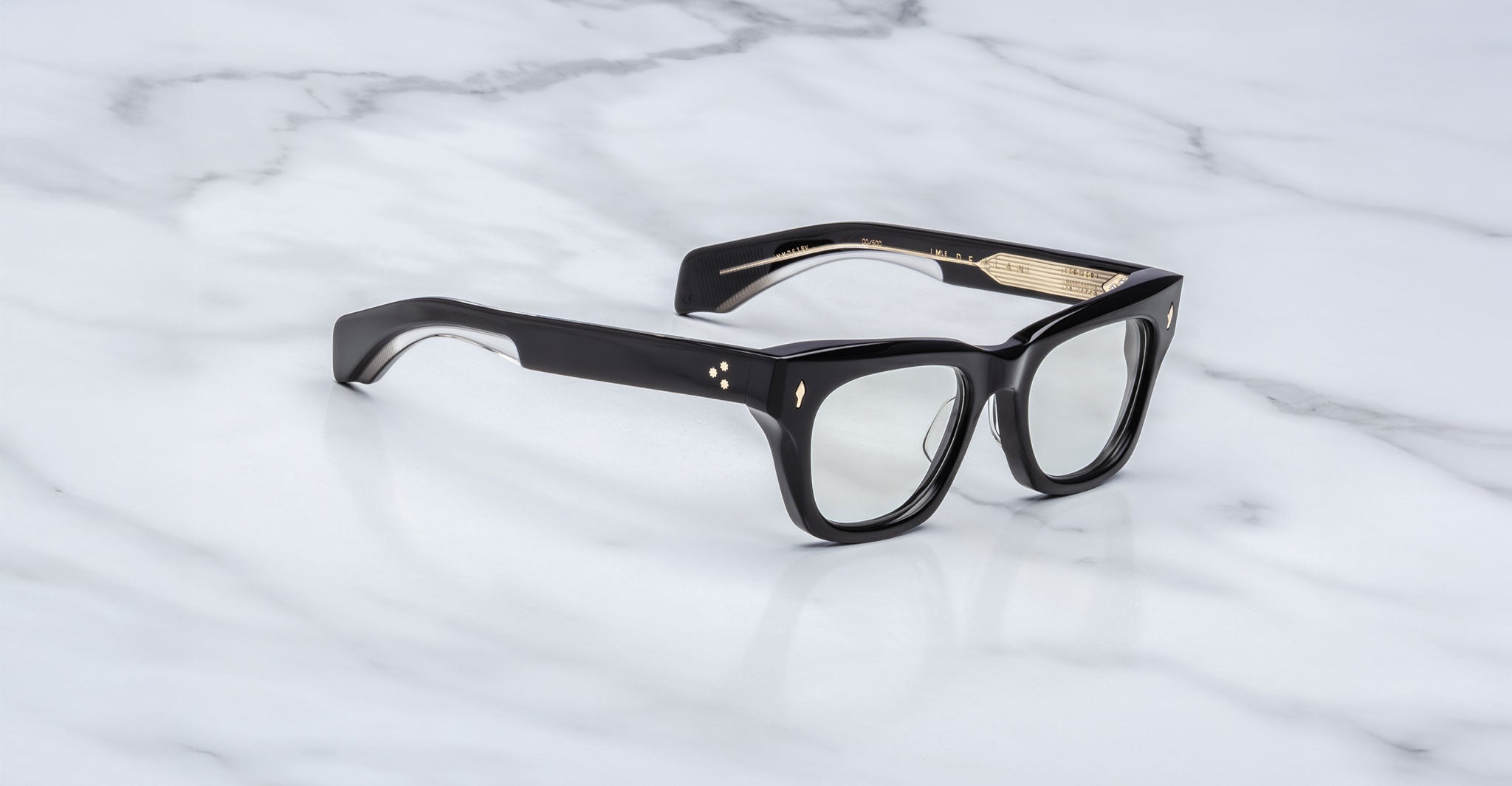 A pair of Jacques Marie Mage Dealan black, thick-rimmed eyeglasses with gold detailing on the inner arms rests on a white marble surface.