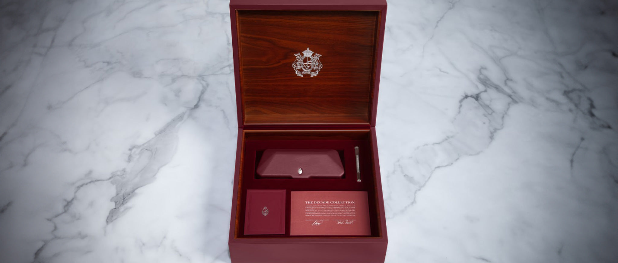 A burgundy wooden box with a crest on the inside lid holds an envelope, card, pen, and Jacques Marie Mage Jagger aviator sunglasses by Jacques Marie Mage, elegantly arranged on a marble surface.