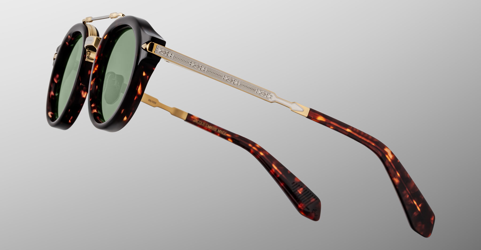 A close-up of Jacques Marie Mage Delage tortoiseshell sunglasses with green lenses, gold engraved metal arms, and a brown and amber mottled frame and temple tips, set against a gray gradient background.