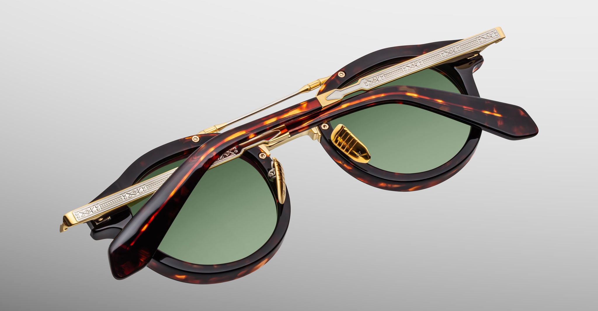A pair of Jacques Marie Mage Delage tortoiseshell sunglasses with green lenses is folded, revealing detailed gold accents and engraved patterns on the arms and bridge against a plain gray background.