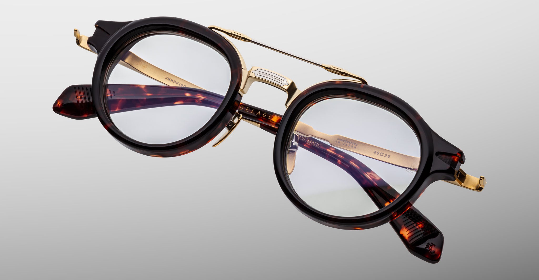 The Jacques Marie Mage Delage is a pair of round eyeglasses featuring tortoiseshell acetate frames, gold metal bridge and temples, and clear lenses, shown against a light gray background.
