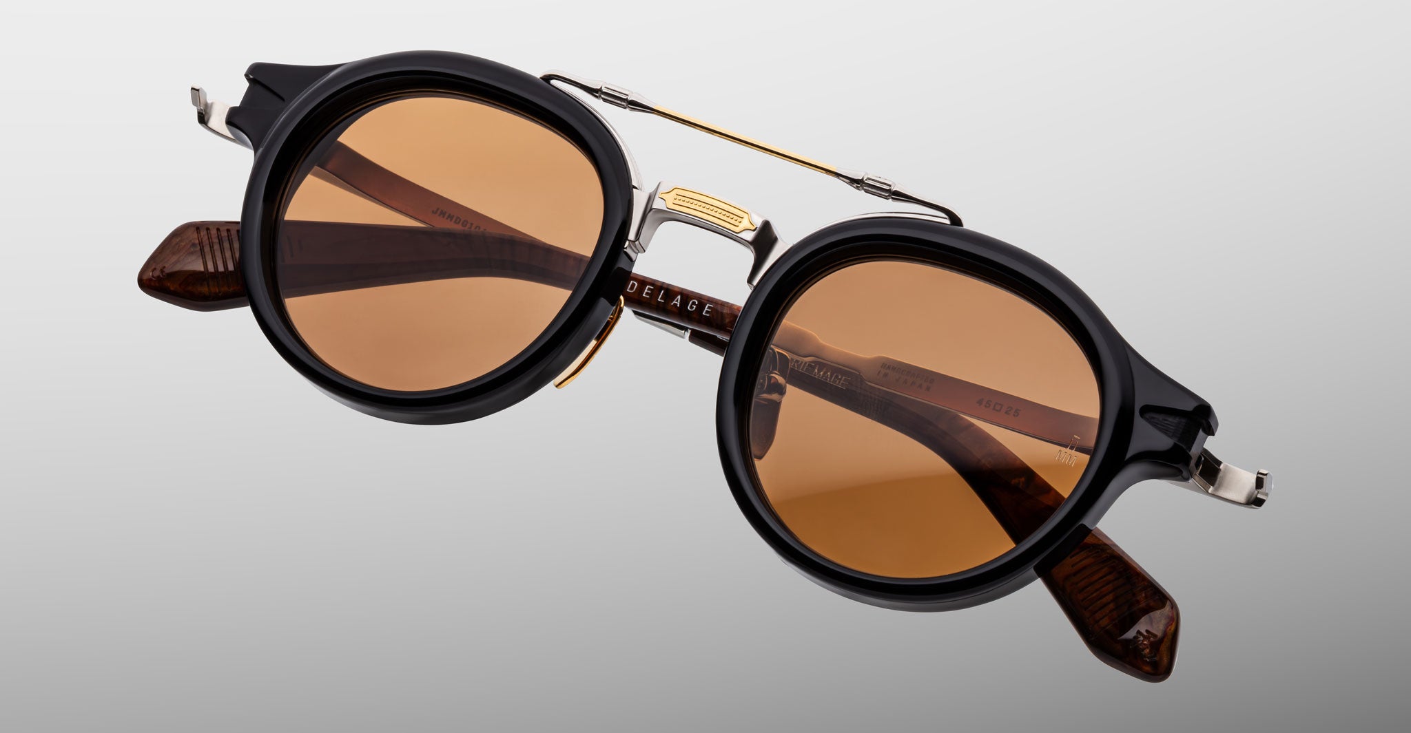 The Jacques Marie Mage Delage is a pair of round sunglasses with black frames, brown-tinted lenses, and gold detailing, featuring metallic accents on the temples and bridge, displayed against a plain gray background.
