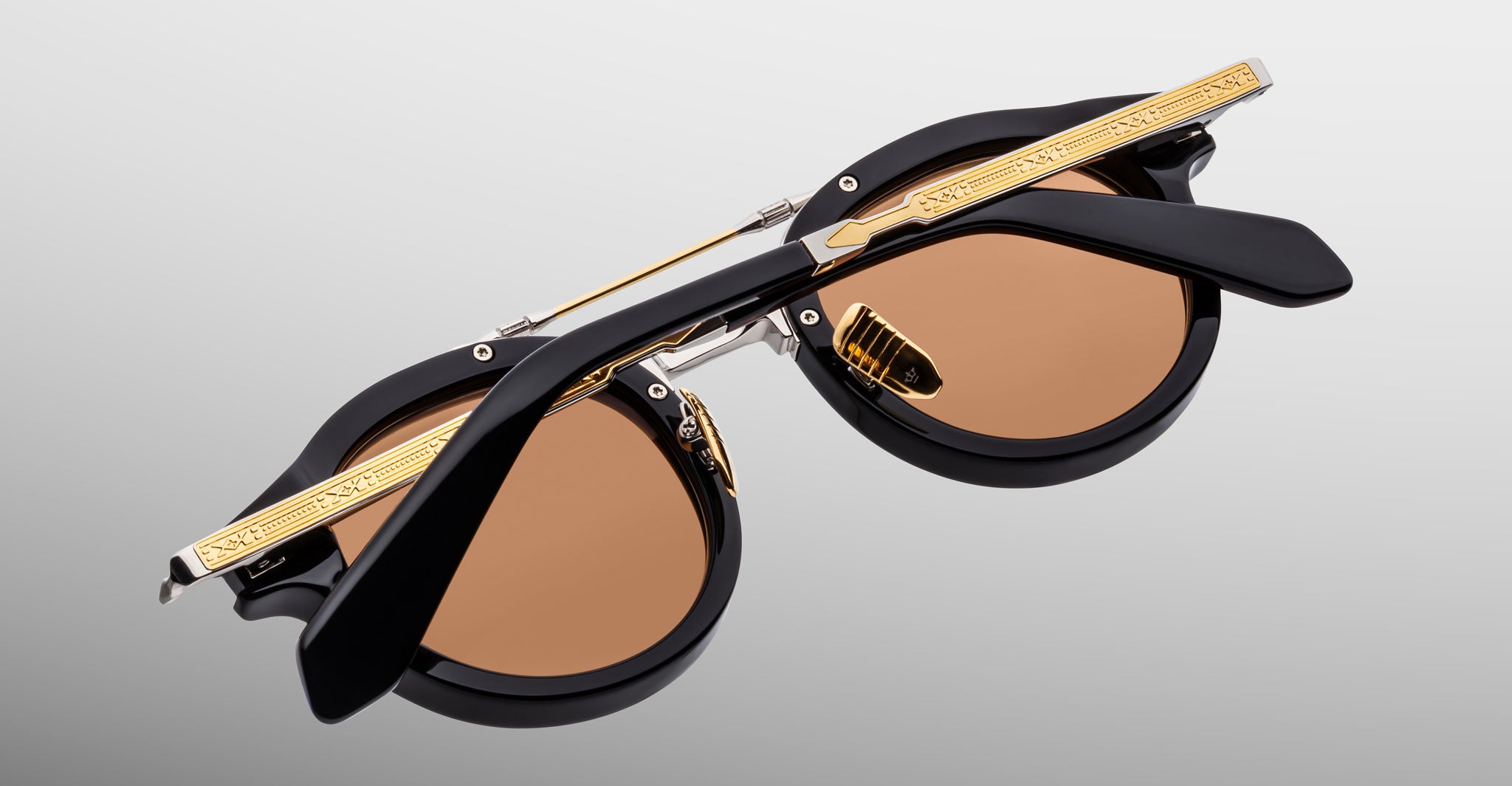 A close-up of Jacques Marie Mage Delage sunglasses featuring black frames with gold detailing and brown lenses, shown folded with crossed arms on a plain light gray background.