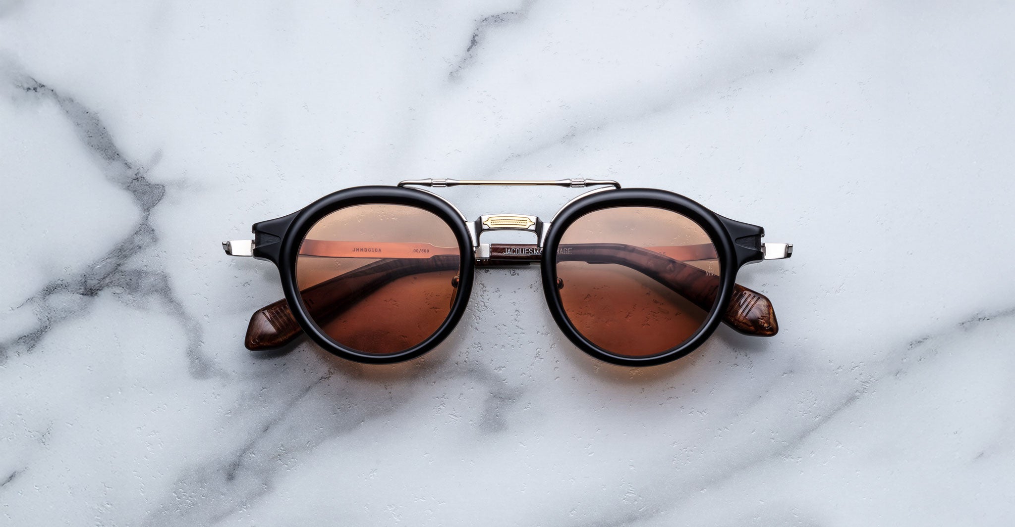 The Jacques Marie Mage Delage sunglasses, featuring a black round frame with brown-tinted lenses and silver accents, rest on a white marble surface.