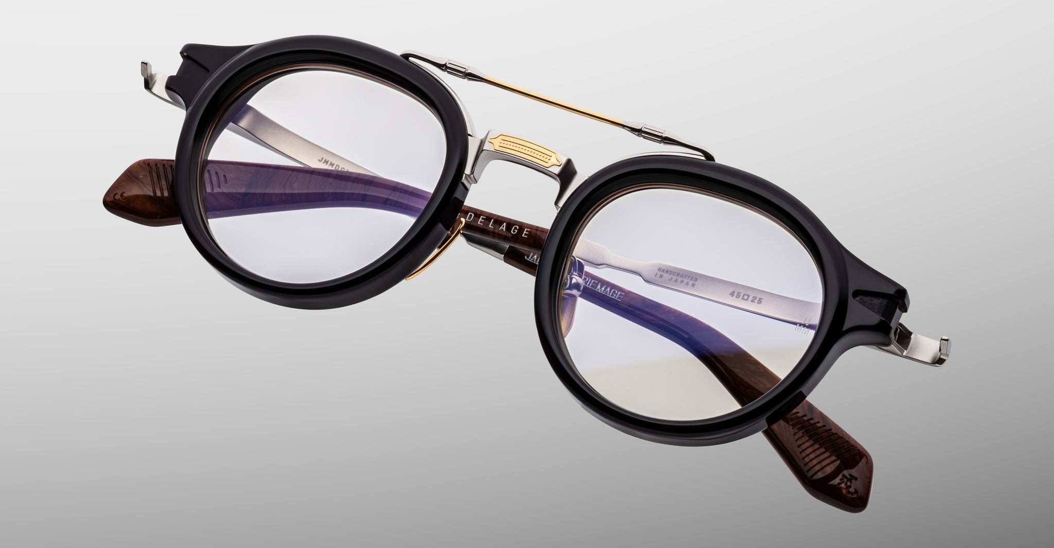 The Jacques Marie Mage Delage eyeglasses feature round black rims, a gold bridge, and brown tortoiseshell temple arms. Sturdy frames with clear prescription lenses are displayed against a light gray background.