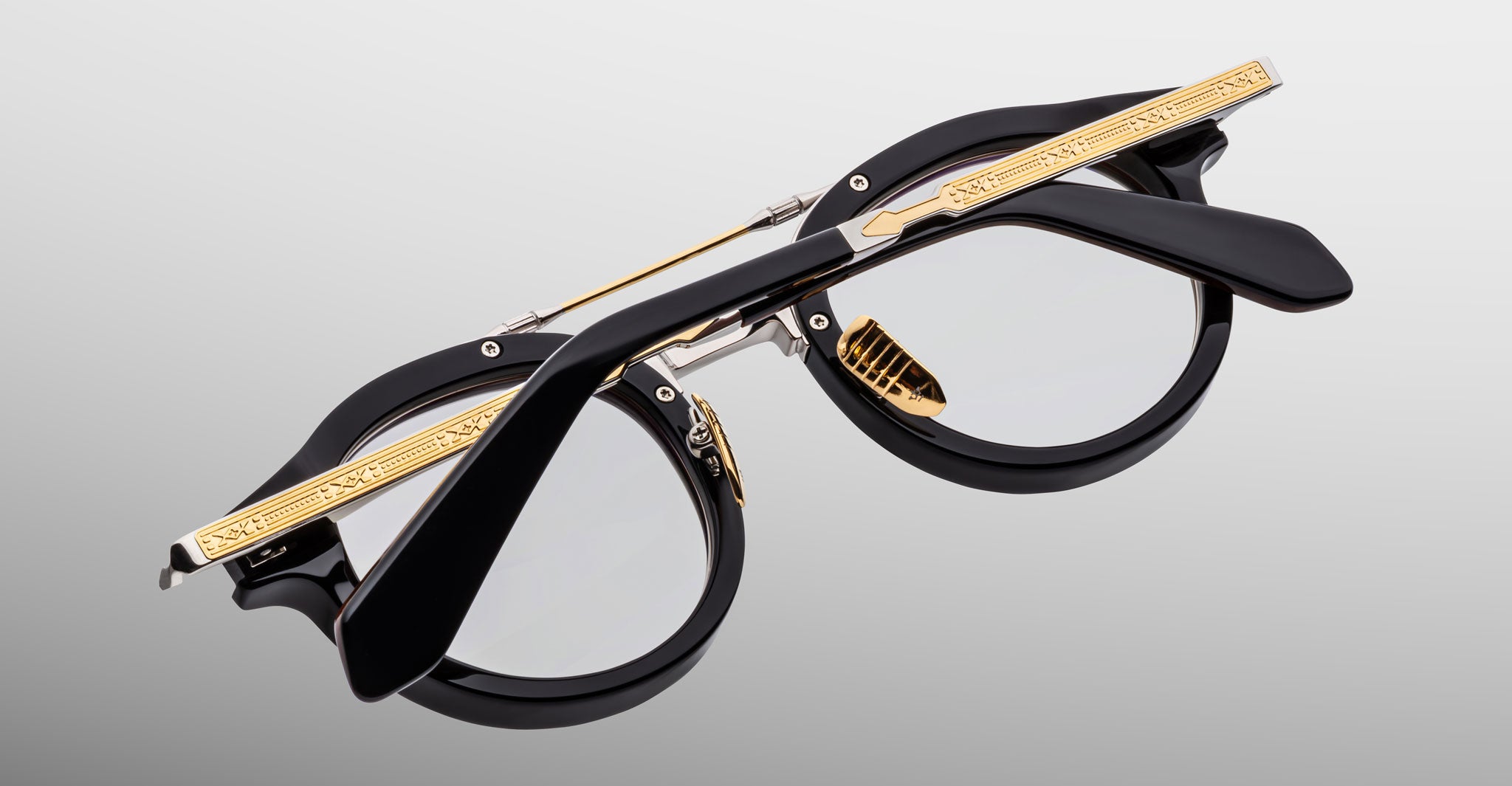 The Jacques Marie Mage Delage: black round eyeglasses with clear lenses, featuring gold detailing on the arms and nose bridge, displayed folded against a light gray background.