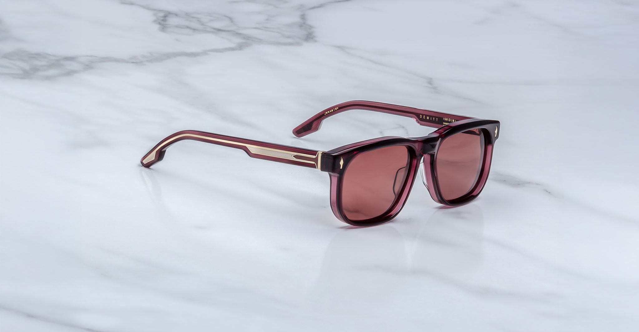 The Jacques Marie Mage Dewitt sunglasses feature maroon rectangular lenses and gold-accented arms, displayed on a white marble surface.