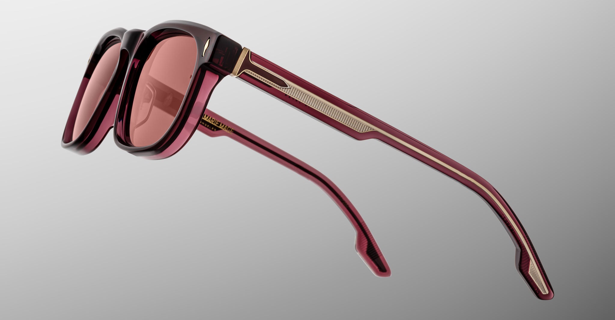 The Jacques Marie Mage Dewitt sunglasses feature a maroon angular frame, tinted lenses, and gold temple detailing, displayed on a light gray background.
