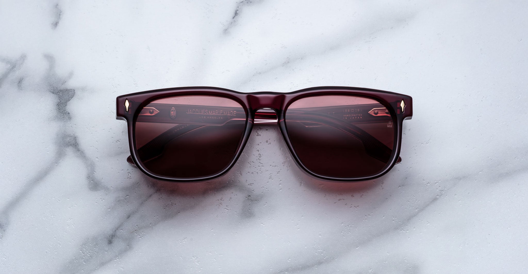 The Jacques Marie Mage Dewitt, square-shaped sunglasses with dark red frames and tinted lenses, are displayed on a white marble surface.