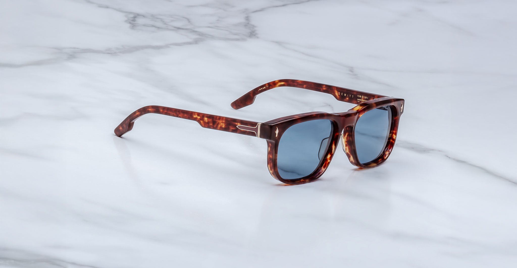 The Jacques Marie Mage Dewitt sunglasses, featuring brown tortoiseshell frames and blue-tinted lenses, rest on a white marble surface.