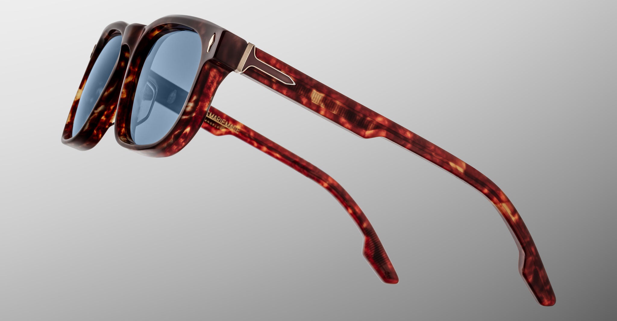 Jacques Marie Mage Dewitt sunglasses in a tortoiseshell pattern with blue lenses, glossy frames, and gold hinge detailing are shown from a side angle against a plain gray background.