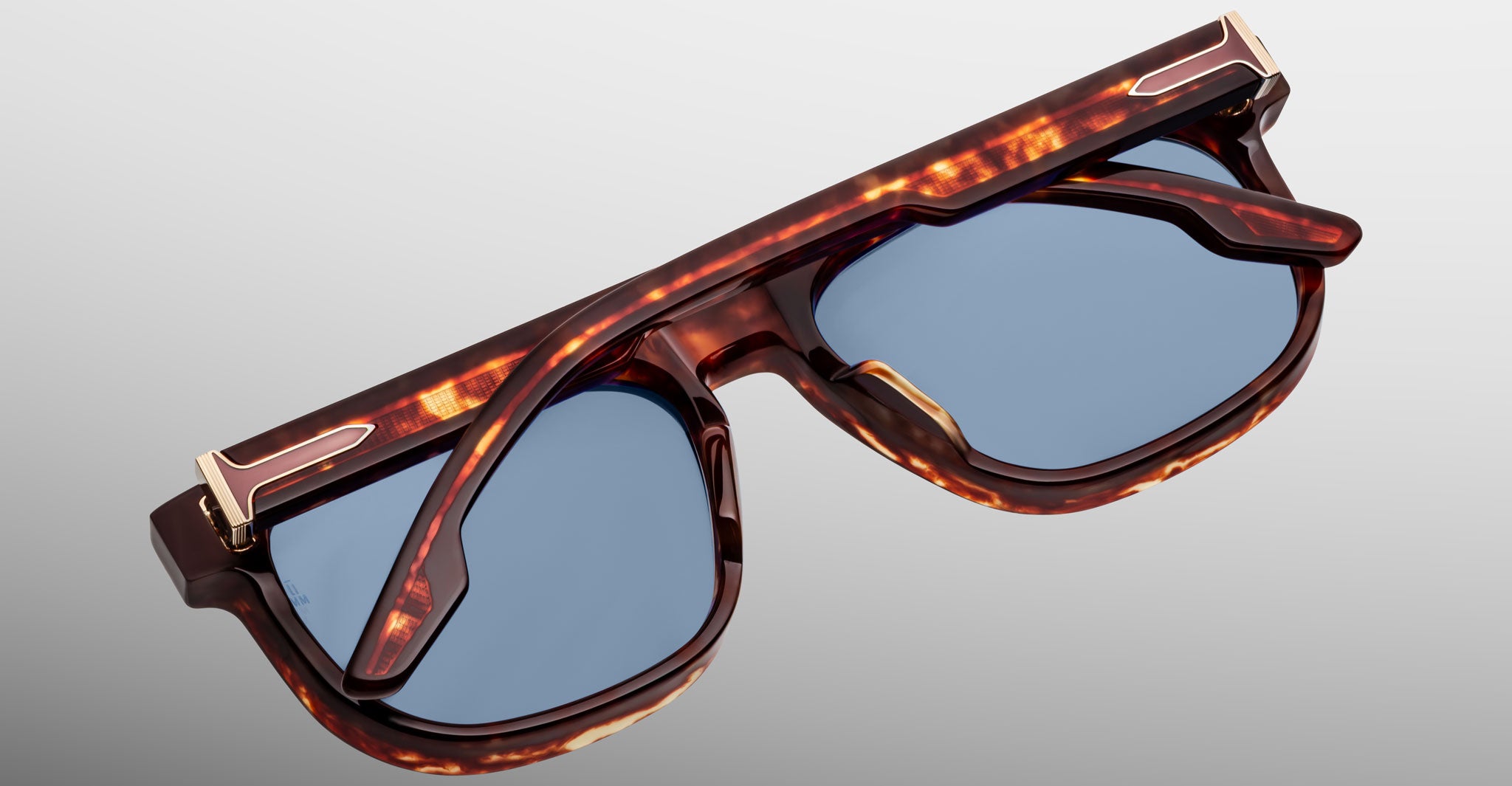 The Jacques Marie Mage Dewitt sunglasses, featuring a tortoiseshell frame with gold corner accents, blue-tinted lenses, and unique angular details, are folded and placed on a light grey surface.