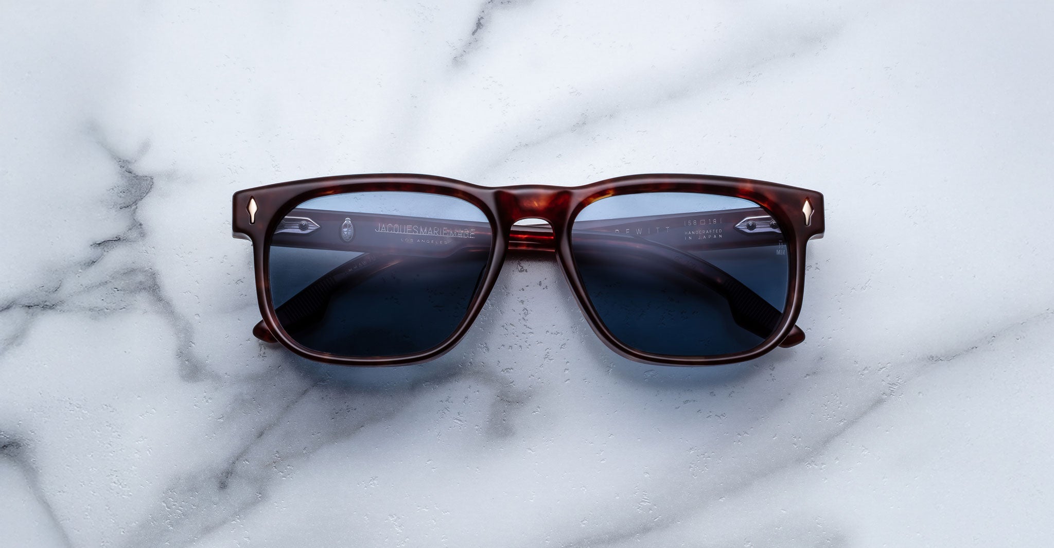 A pair of Jacques Marie Mage Dewitt tortoiseshell sunglasses with dark lenses rests on a white marble surface with gray veining. The arms are slightly open, and the lenses catch some light.